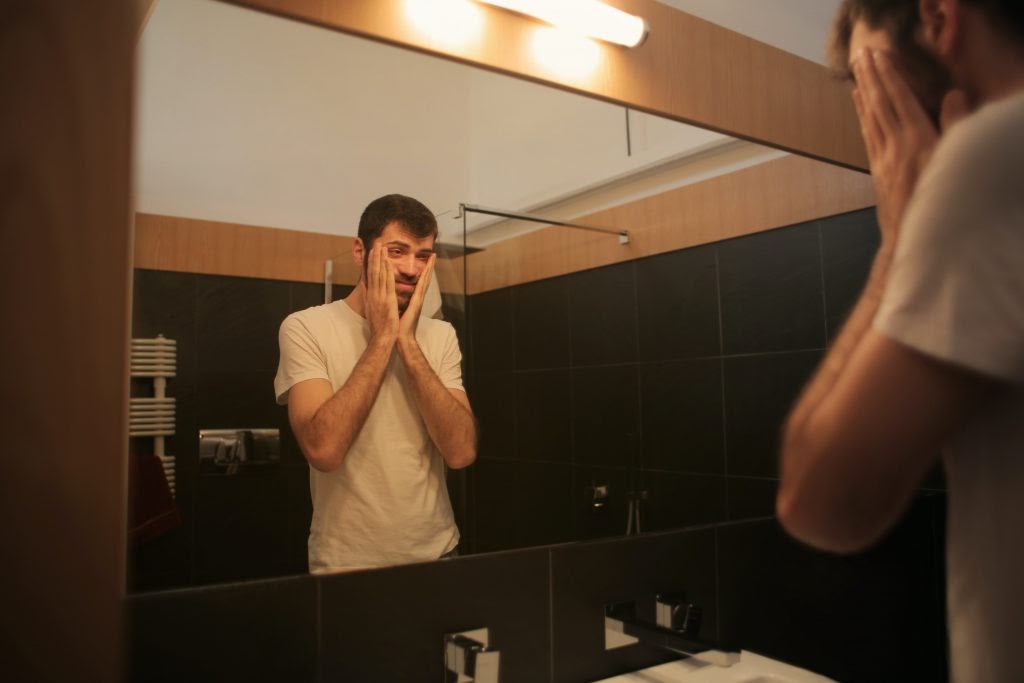 Homem leva as mão ao rosto olhando no espelho do banheiro.